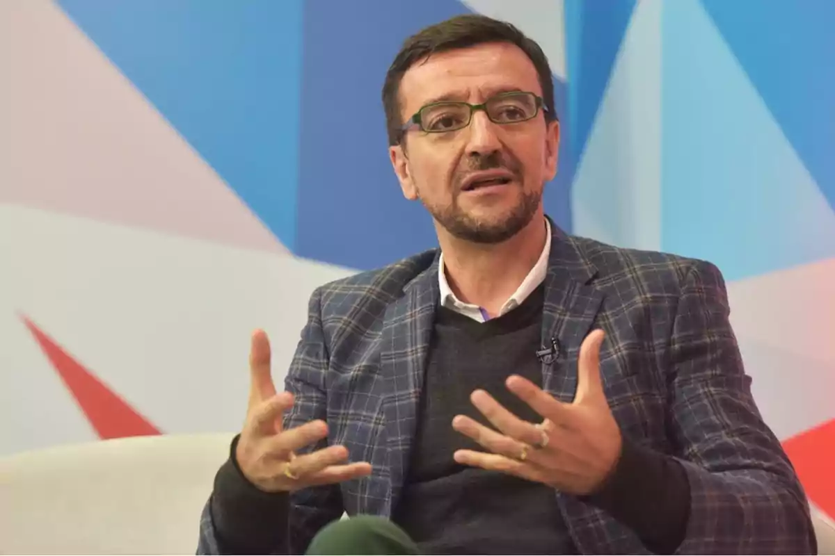 Un hombre con gafas y barba hablando gesticulando con las manos en un entorno con fondo geométrico azul y rojo.