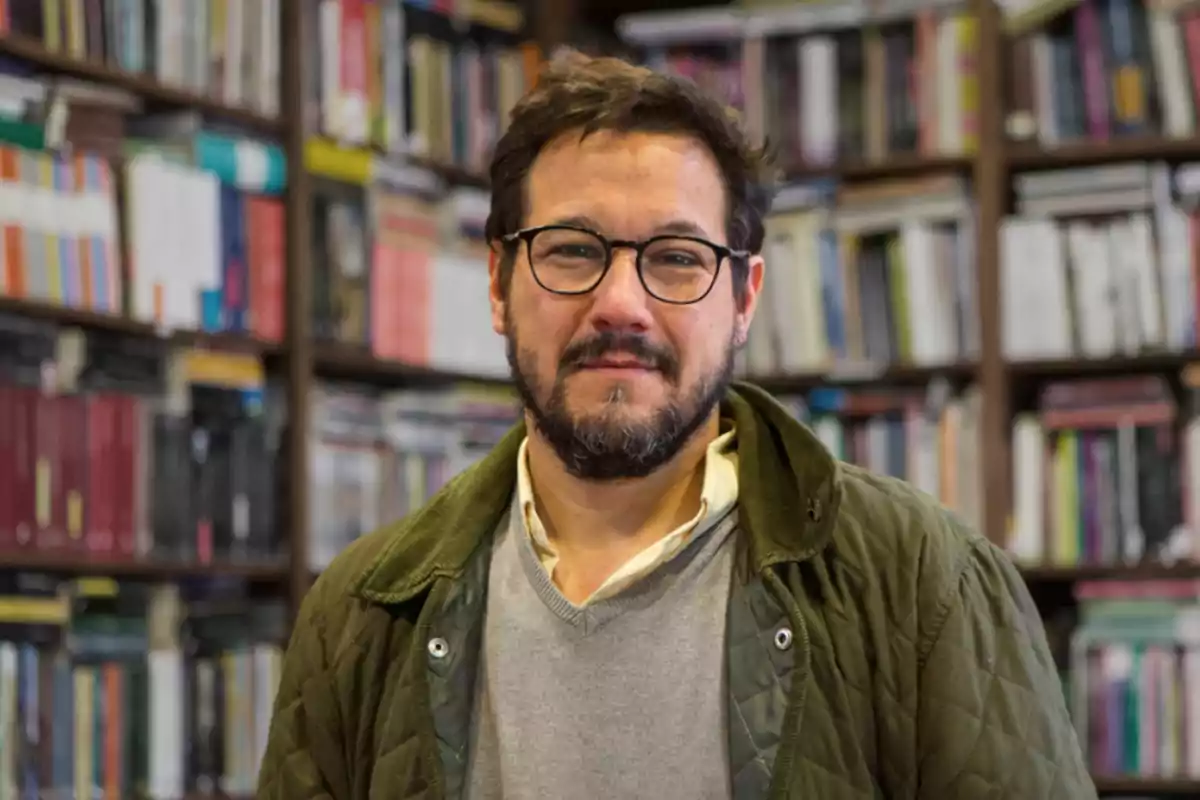 Hombre con gafas y barba en una biblioteca con estanterías llenas de libros.