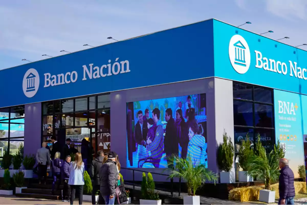 Fachada de una sucursal del Banco Nación con personas entrando y un gran cartel publicitario en el exterior.