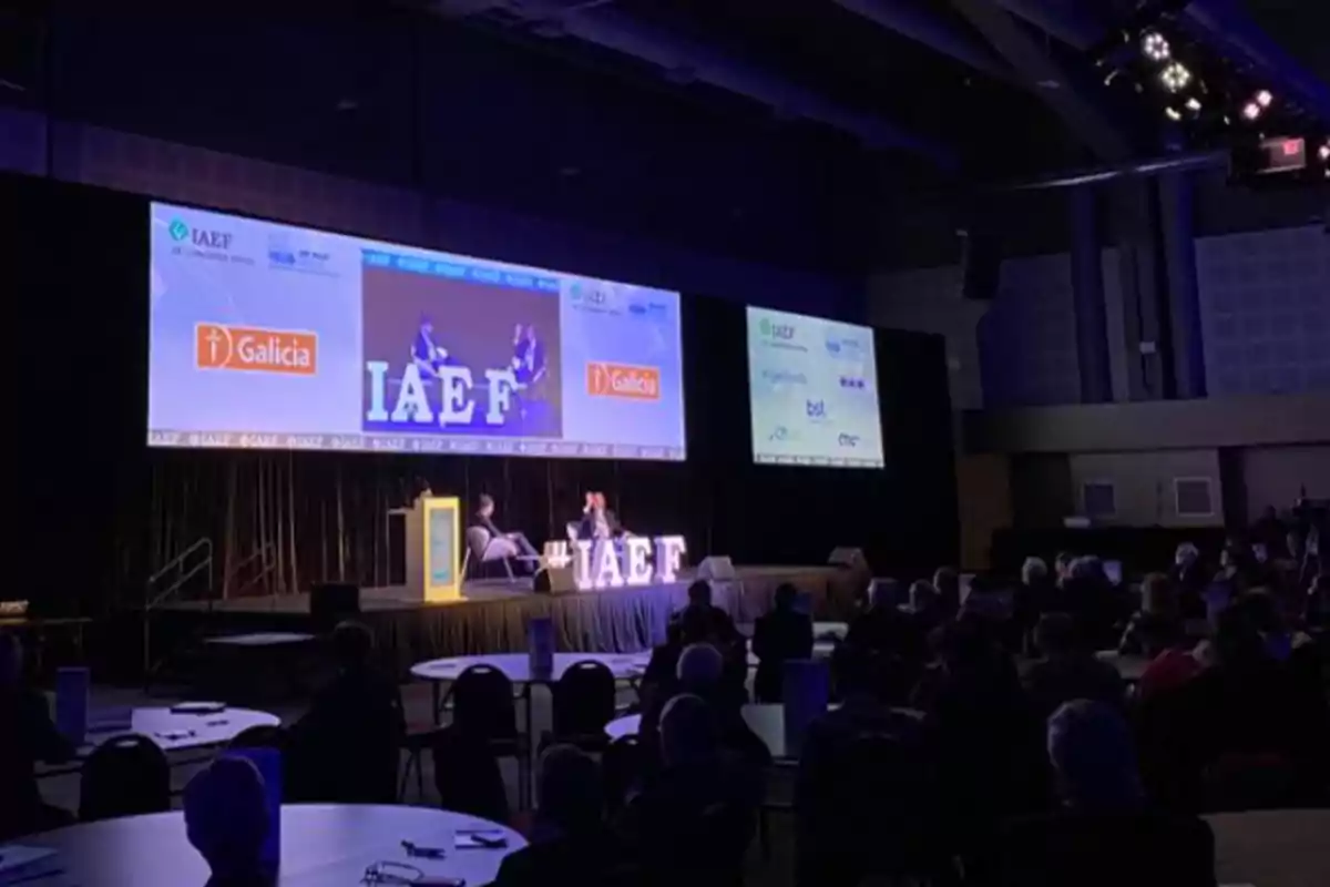 Una conferencia en un auditorio con un escenario iluminado y pantallas grandes mostrando logotipos y el texto IAEF, con personas sentadas en mesas redondas.