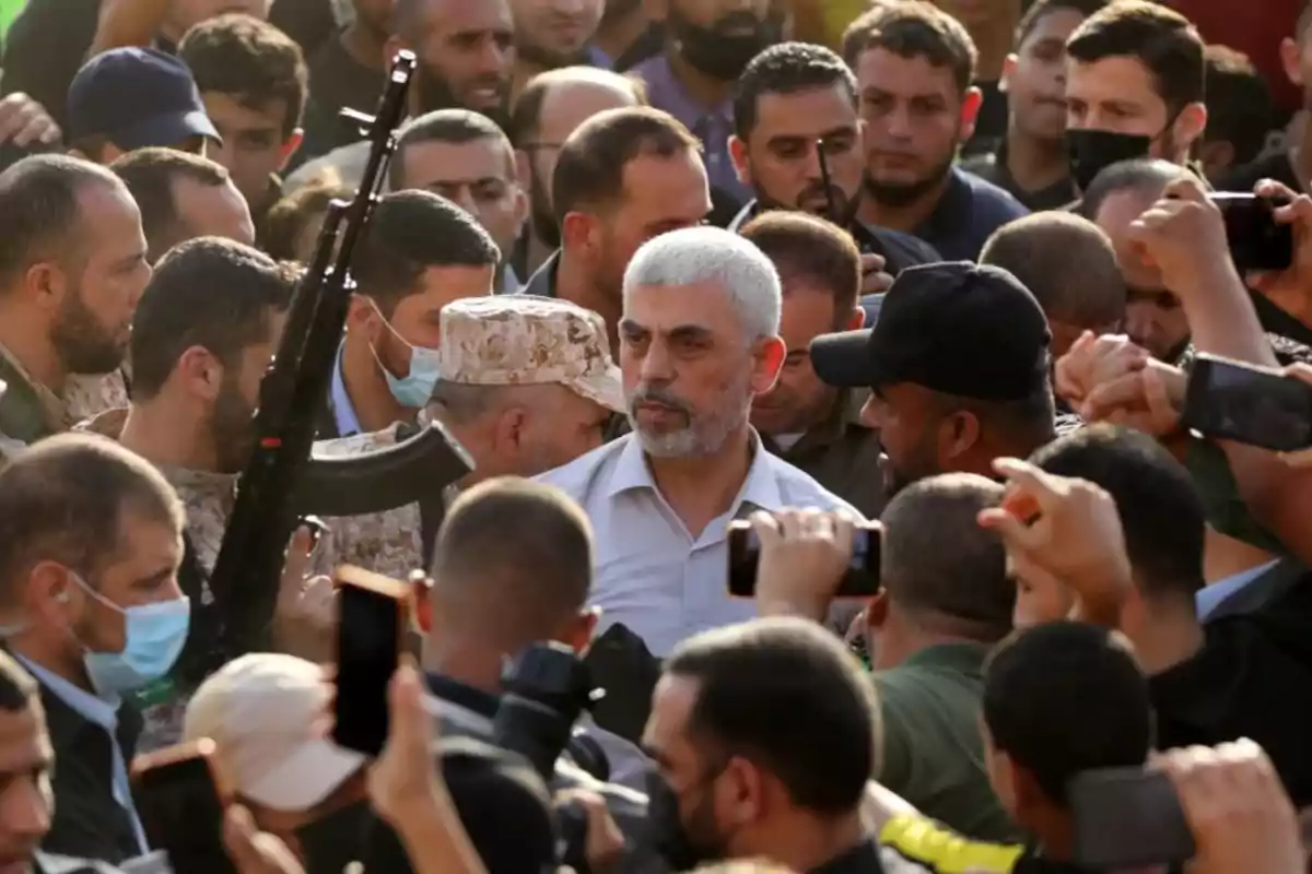 Un hombre de cabello canoso rodeado de una multitud de personas, algunas de las cuales llevan uniformes militares y sostienen cámaras y teléfonos móviles.