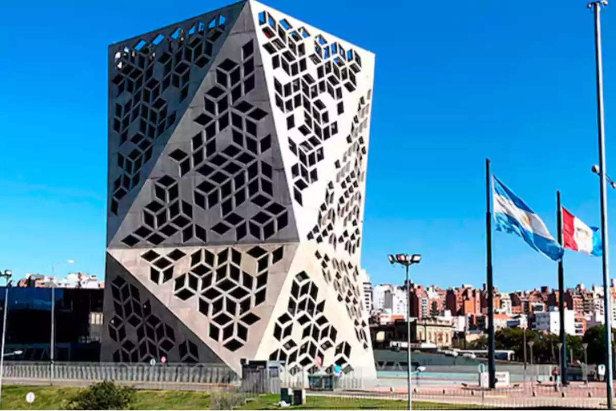 Edificio moderno con diseño geométrico en su fachada junto a dos banderas ondeando en un día soleado.