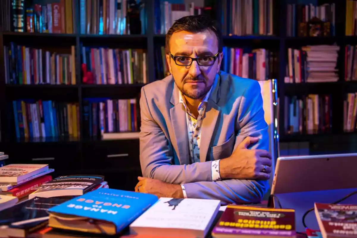 Un hombre con gafas y barba está sentado en un escritorio rodeado de libros, con una estantería llena de más libros al fondo.