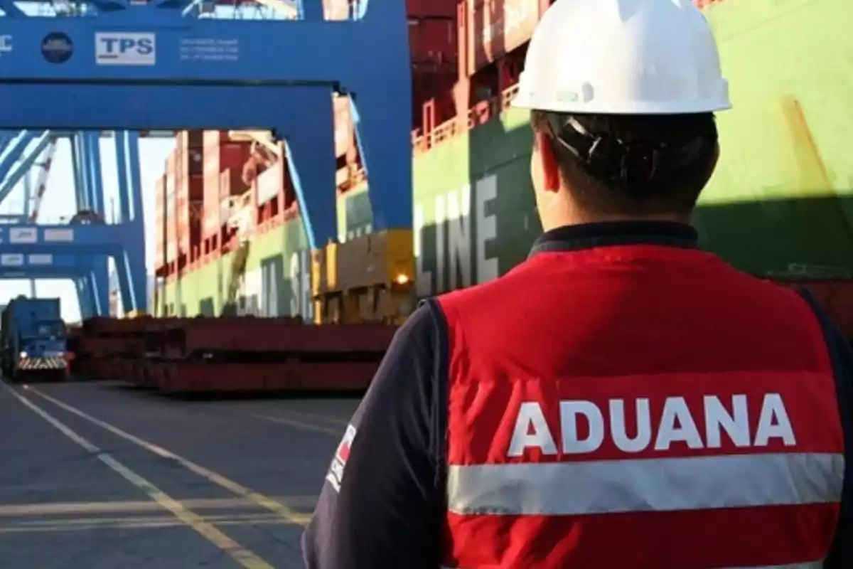 Un trabajador de aduanas con chaleco rojo y casco blanco observa un barco de carga en un puerto.