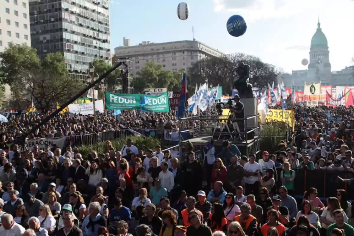 Una multitud se reúne en una manifestación frente a un edificio gubernamental con pancartas y globos en el aire.