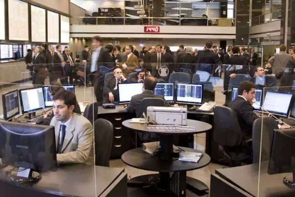 Personas trabajando en una sala de operaciones con múltiples computadoras y pantallas, mientras otras personas caminan y conversan en el fondo.