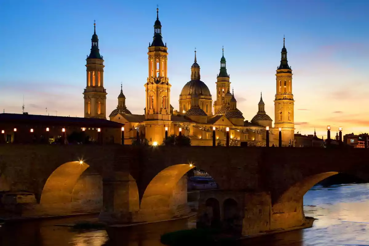 Vista nocturna de una catedral iluminada con un puente de piedra en primer plano y un cielo al atardecer.