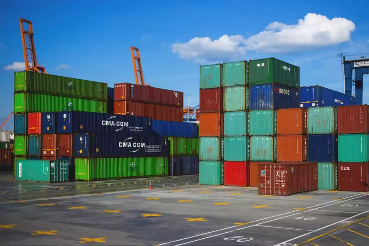 Contenedores de carga apilados en un puerto con grúas al fondo bajo un cielo azul.