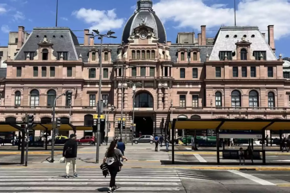 Una estación de tren histórica con una fachada de estilo clásico y un reloj en la parte superior, personas cruzando la calle frente al edificio bajo un cielo azul con algunas nubes.