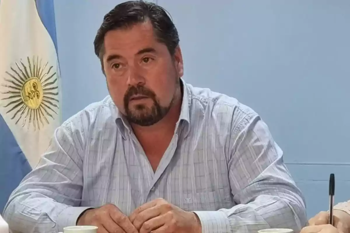 Hombre con barba y camisa a cuadros sentado frente a una bandera de Argentina.