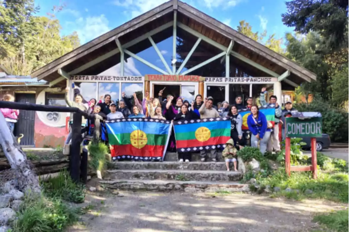 Un grupo de personas sosteniendo banderas mapuches frente a un edificio de madera con letreros que indican un comedor.