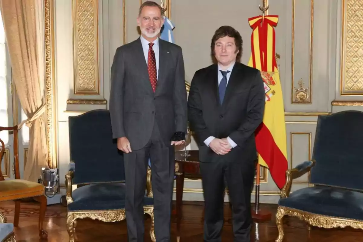 Dos hombres de traje posan en una sala elegante con banderas de Argentina y España al fondo.