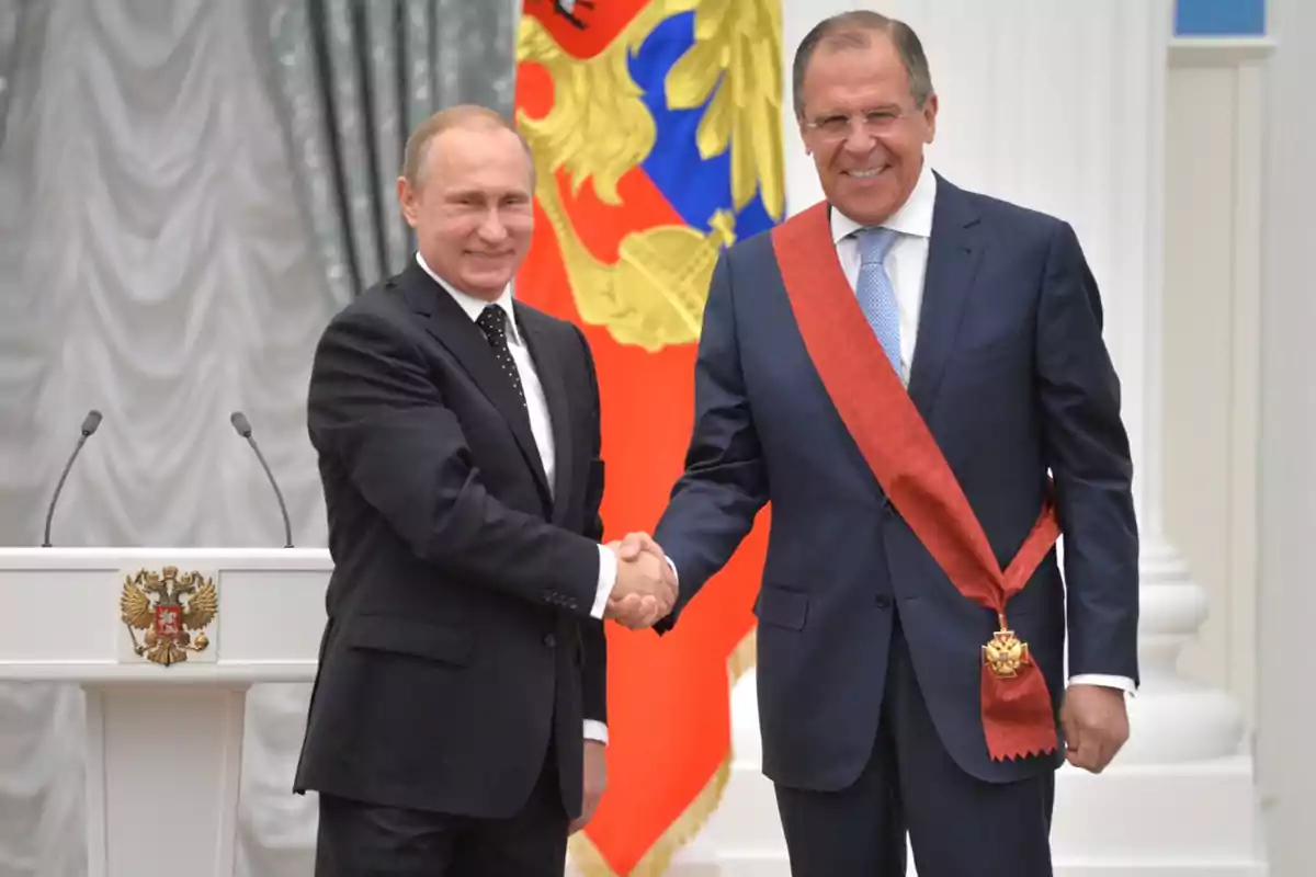 Dos hombres de traje se dan la mano frente a un podio con un escudo y una bandera colorida de fondo.