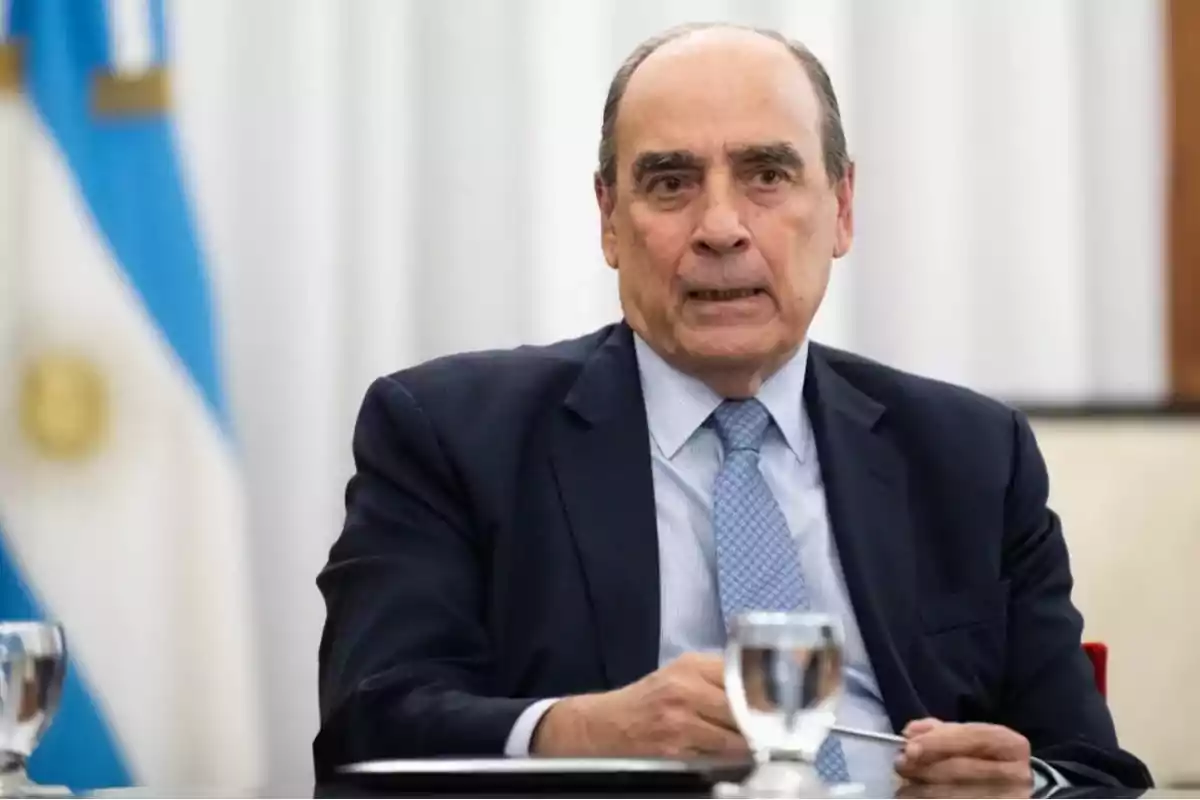 Hombre de traje oscuro y corbata azul claro sentado frente a una bandera argentina.