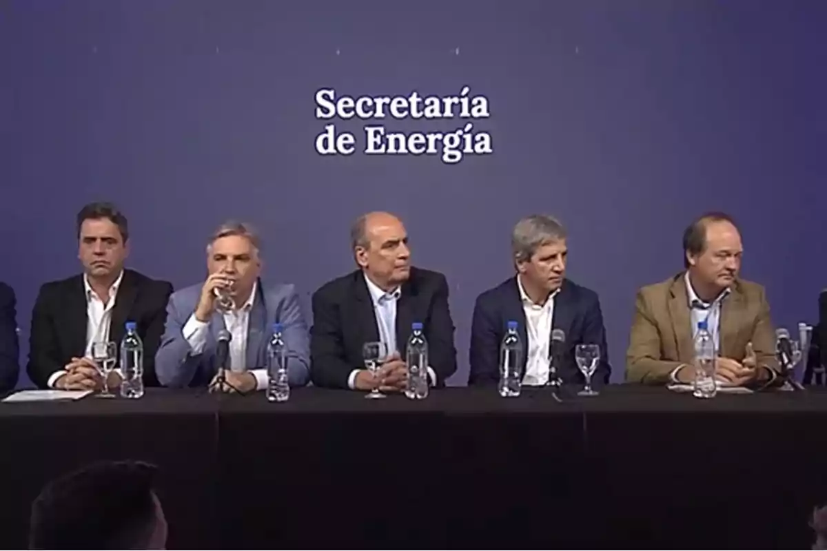 Un grupo de hombres sentados en una mesa con botellas de agua y micrófonos frente a un fondo que dice 