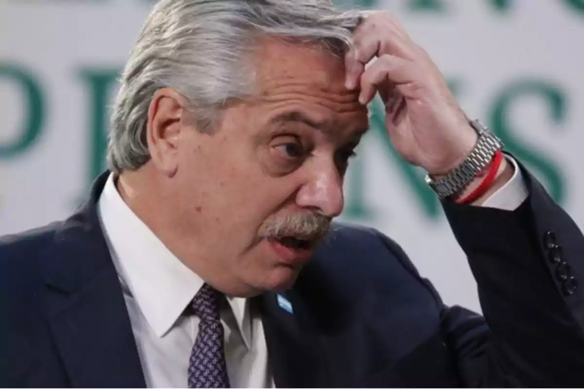 Hombre de cabello canoso y bigote con expresión pensativa tocándose la cabeza vestido con traje y corbata.