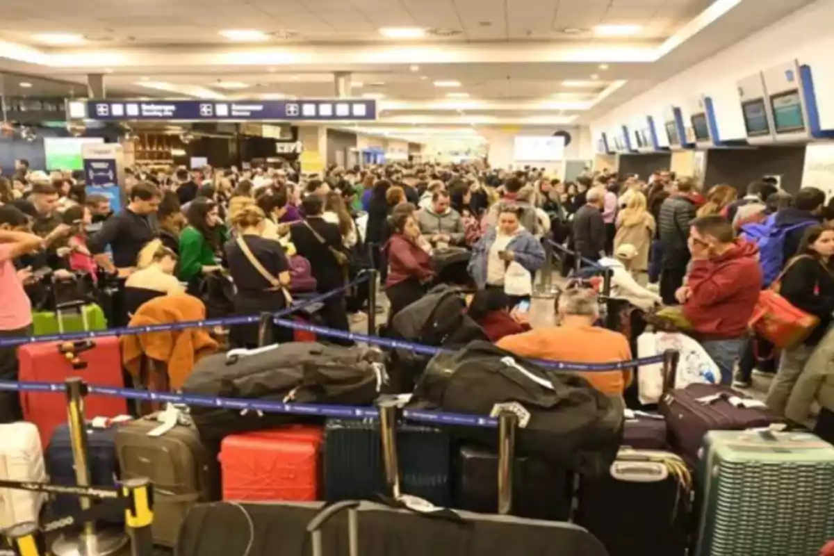 Una multitud de personas esperando en una fila en un aeropuerto con muchas maletas alrededor.