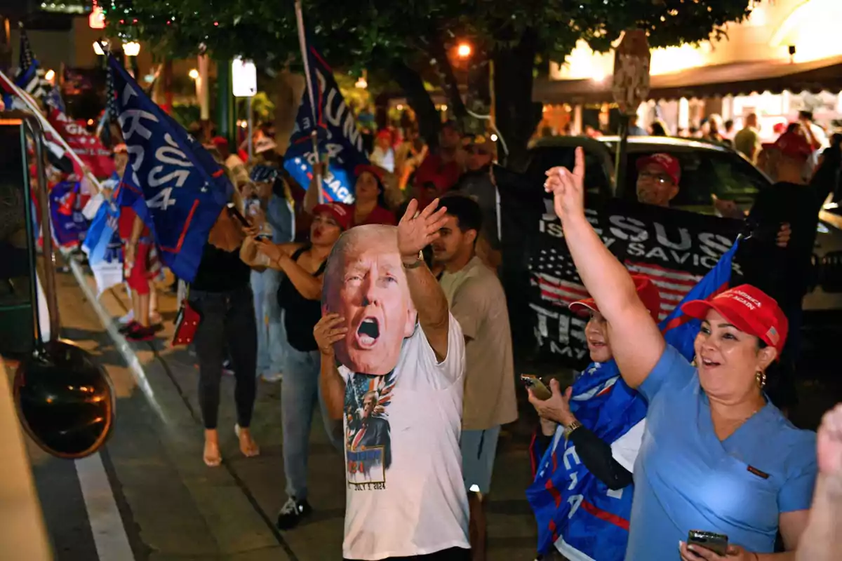 Un grupo de personas participa en una manifestación nocturna, ondeando banderas y portando carteles, algunas con gorras rojas y una máscara de una figura pública.