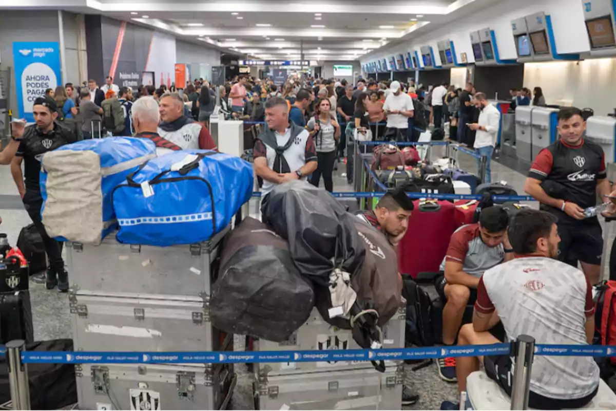 Un grupo de personas con equipaje en un aeropuerto concurrido, con mostradores de facturación al fondo.