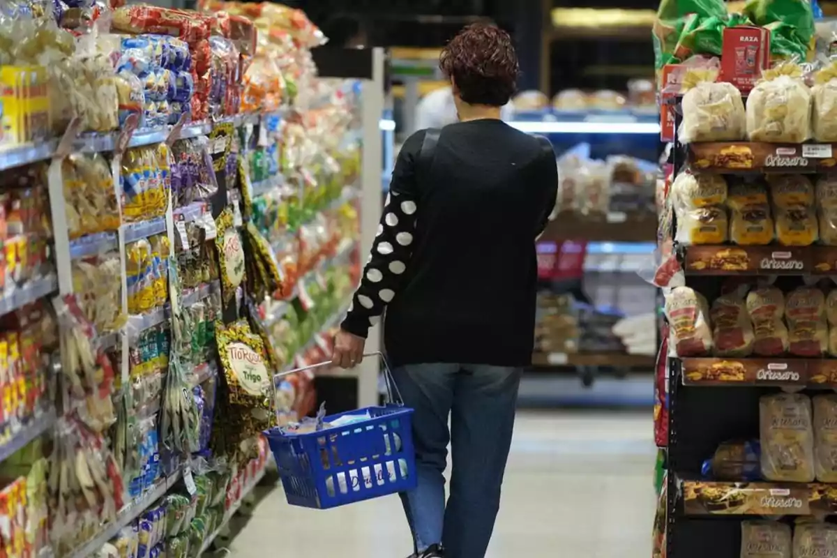 Persona comprando en un supermercado con un carrito azul en el pasillo de productos empaquetados.