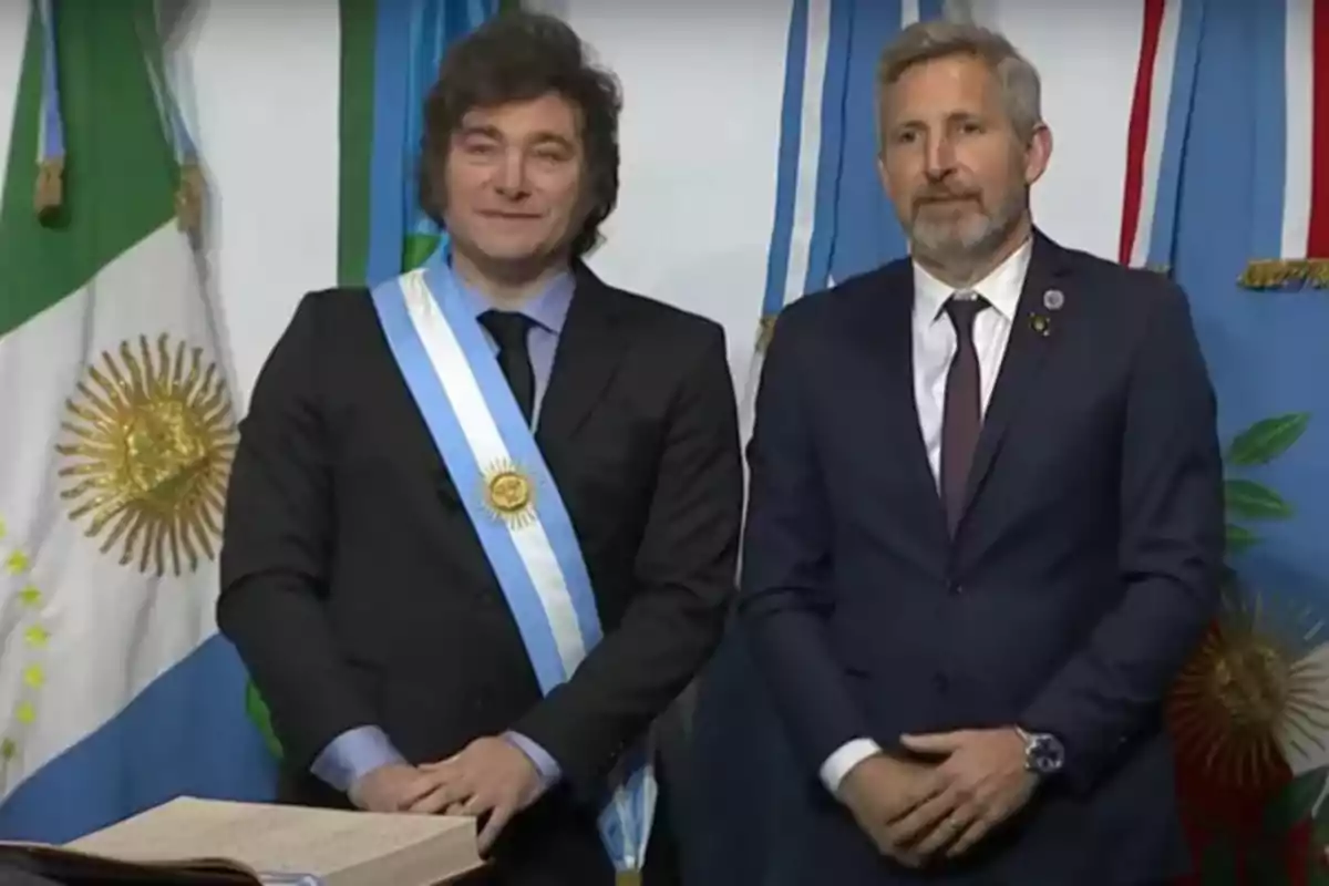 Dos hombres de pie con trajes formales y una banda presidencial, frente a banderas argentinas.