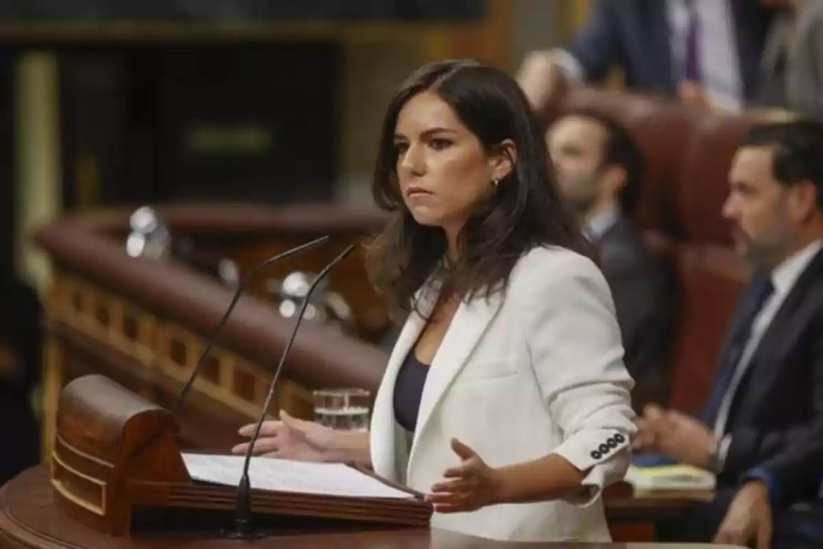 Mujer de cabello oscuro hablando en un podio en un entorno formal con personas sentadas al fondo.