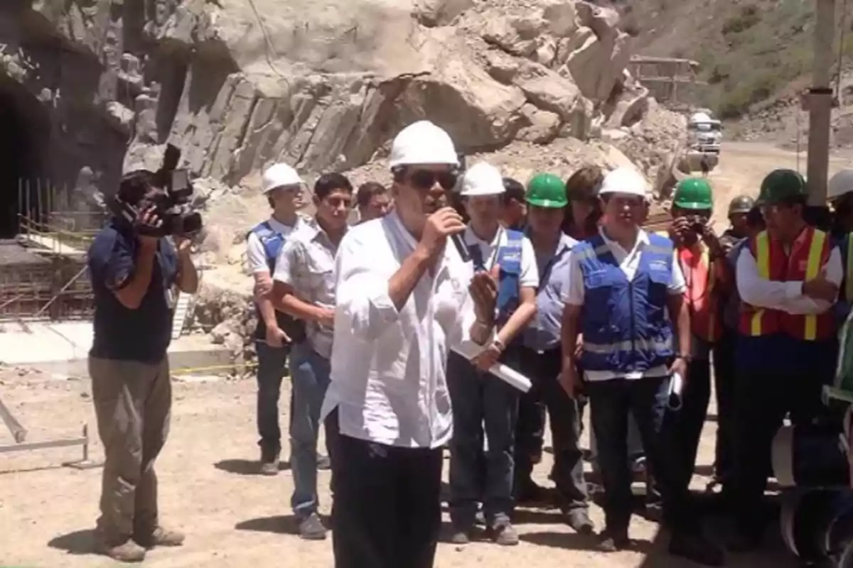 Un grupo de personas con cascos y chalecos de seguridad se encuentra en un sitio de construcción al aire libre mientras un hombre habla por un micrófono y un camarógrafo graba la escena.