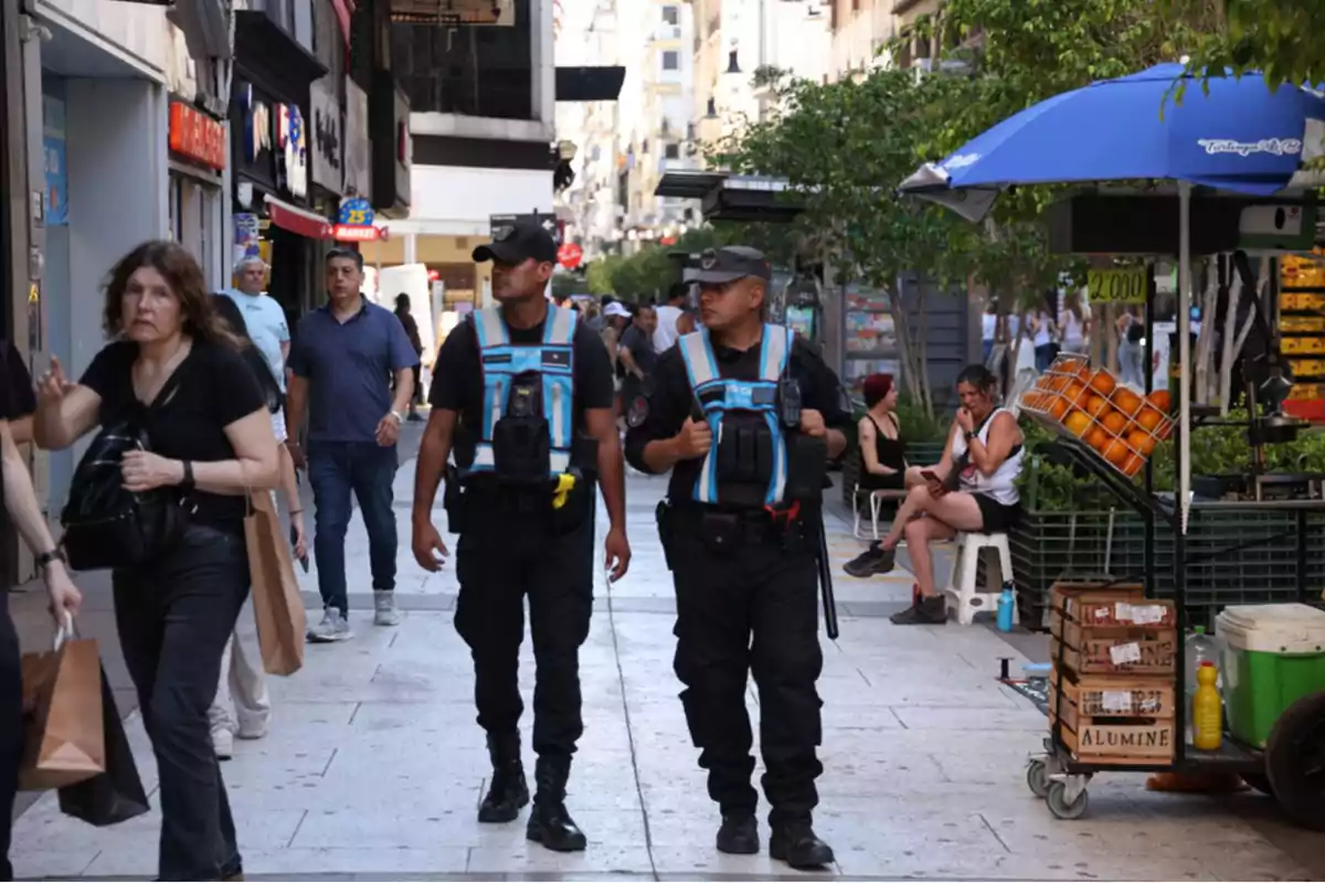 Dos policías patrullan una calle concurrida con tiendas y un puesto de frutas bajo una sombrilla azul.