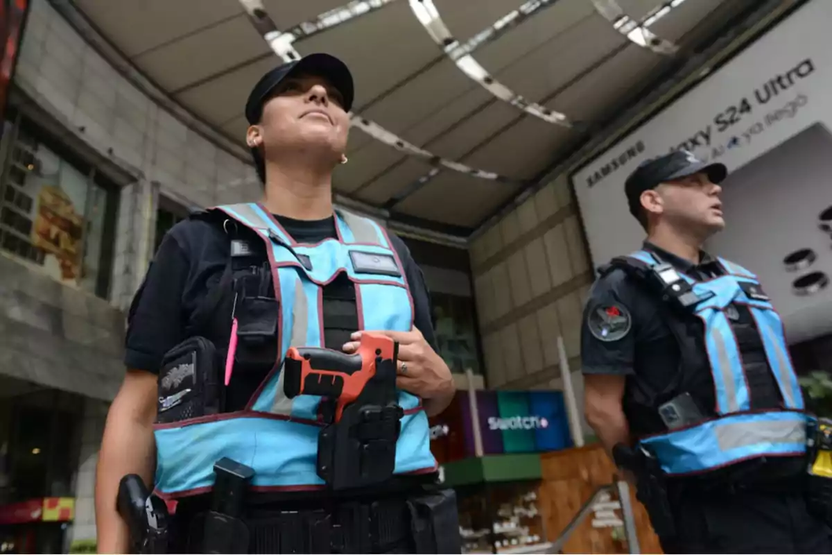 Dos guardias de seguridad con chalecos azules y negros están de pie en el interior de un centro comercial.