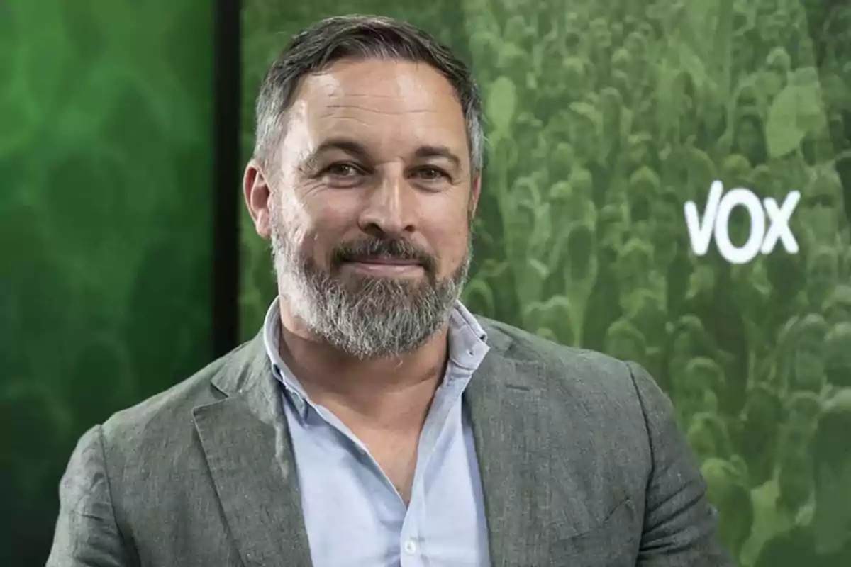 Un hombre con barba y cabello corto, vestido con camisa azul y chaqueta gris, frente a un fondo verde con el logo de Vox.