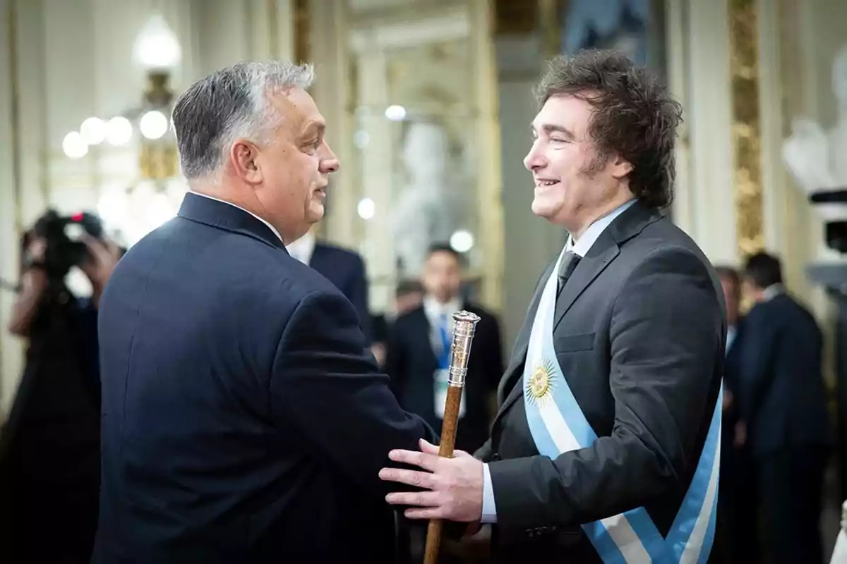 Dos hombres de traje se saludan cordialmente en un entorno formal con decoración elegante y personas al fondo.