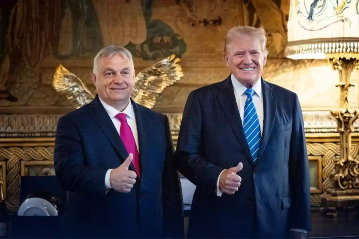 Dos hombres de traje sonríen y levantan el pulgar en un entorno elegante.