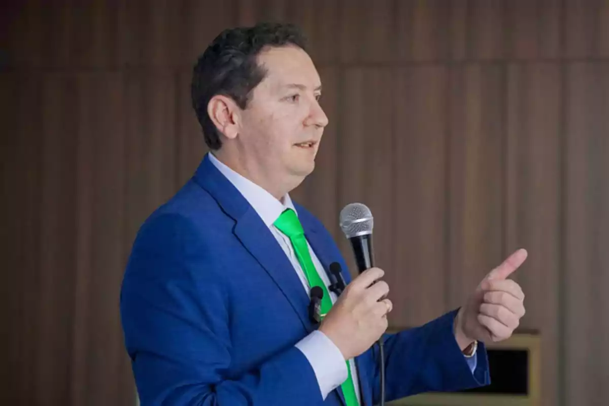 Hombre con traje azul y corbata verde hablando en público mientras sostiene un micrófono.