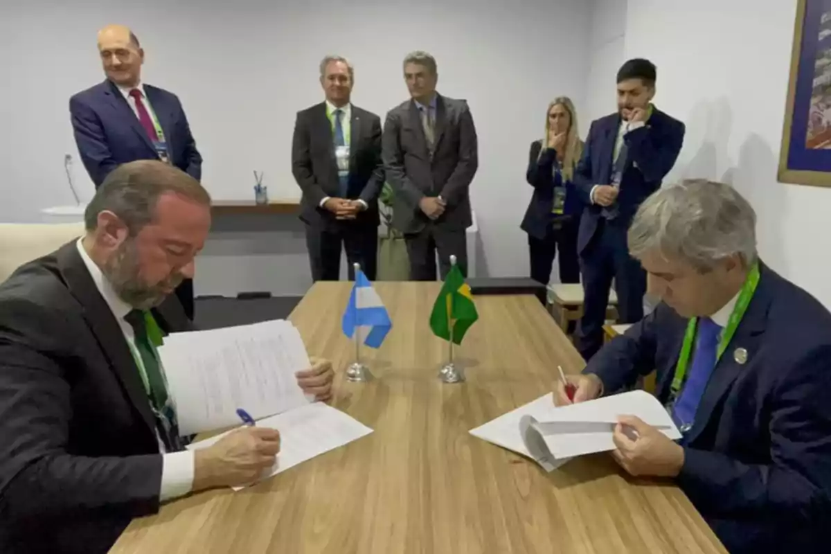 Dos hombres firmando documentos en una mesa con banderas de Argentina y Brasil, mientras otras personas observan de pie en el fondo.