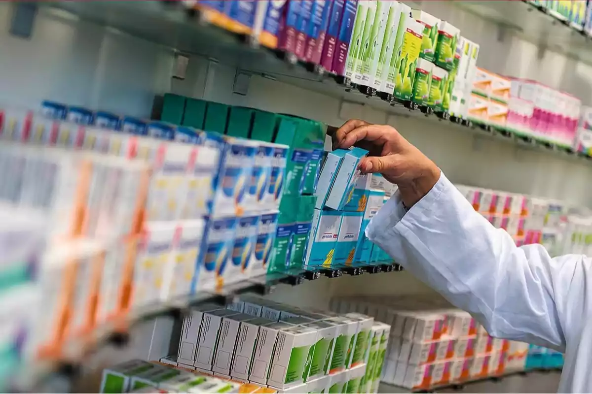 Persona con bata blanca eligiendo una caja de medicamentos en un estante de farmacia.