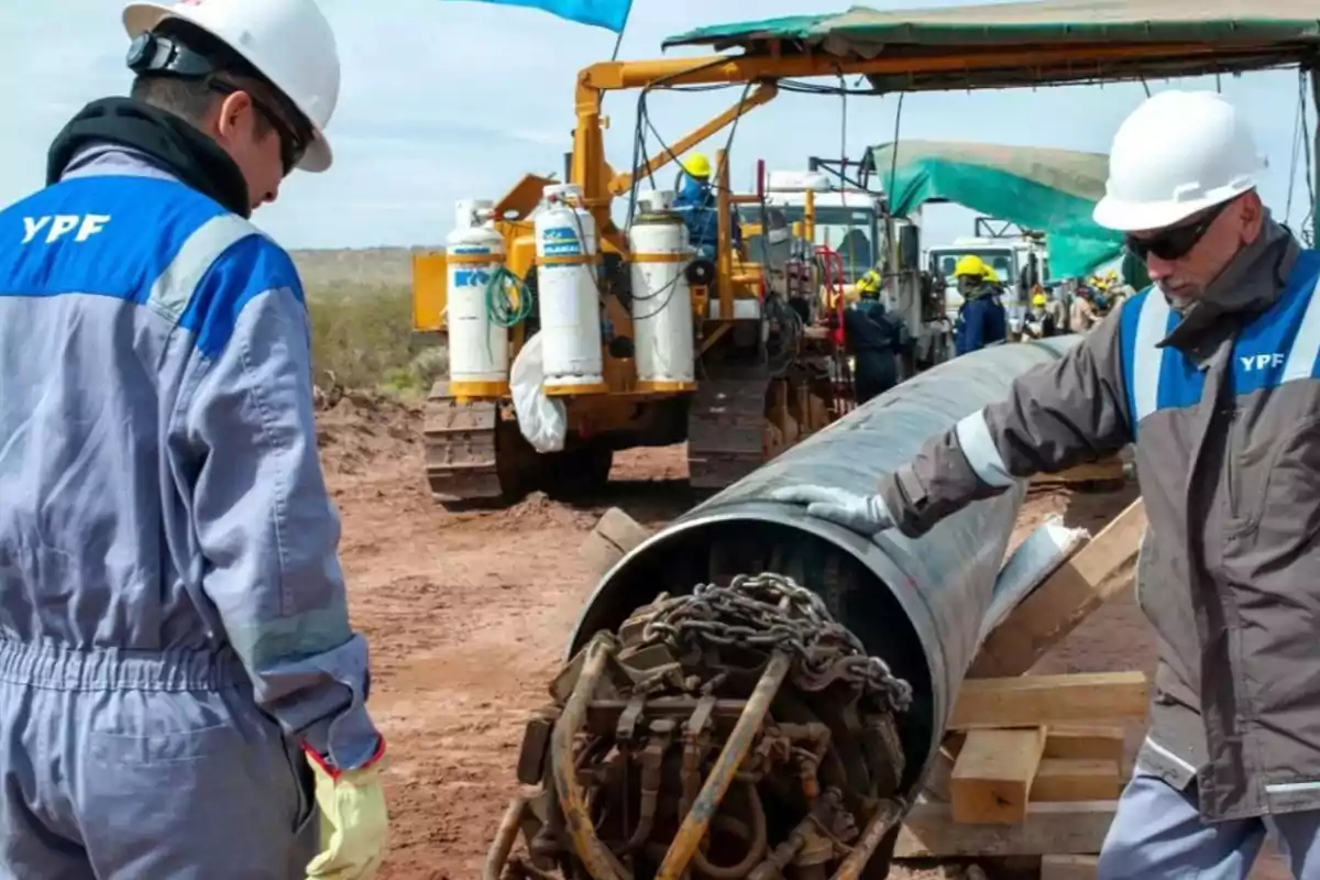 Dos trabajadores con cascos y uniformes de YPF inspeccionan un gran tubo de metal en un sitio de construcción al aire libre, con maquinaria pesada y otros trabajadores al fondo.