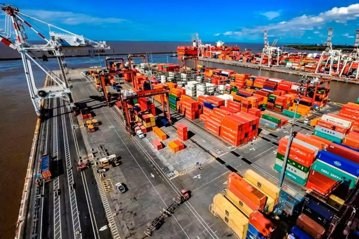 Vista aérea de un puerto con numerosos contenedores de carga apilados y grúas en funcionamiento junto al agua bajo un cielo azul.