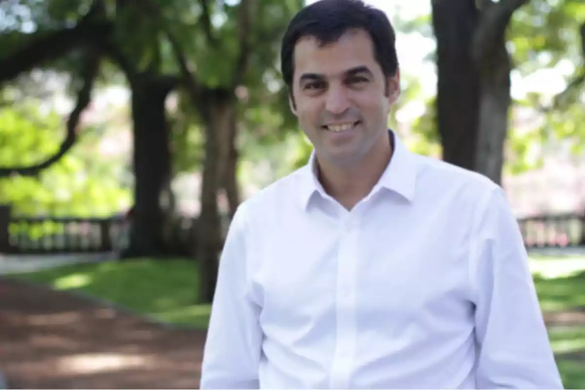 Hombre con camisa blanca sonriendo en un parque con árboles al fondo.