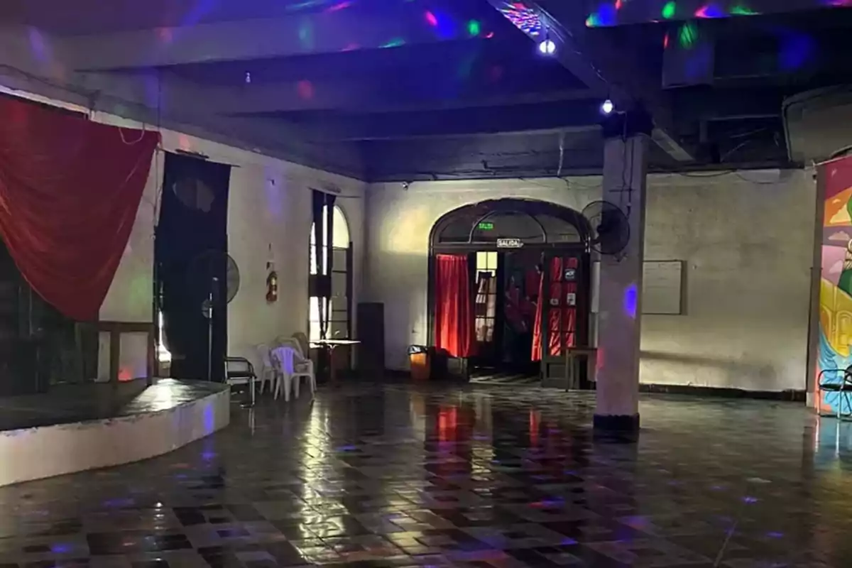 Un salón amplio con piso de baldosas, luces de colores en el techo, cortinas rojas y sillas de plástico blancas.