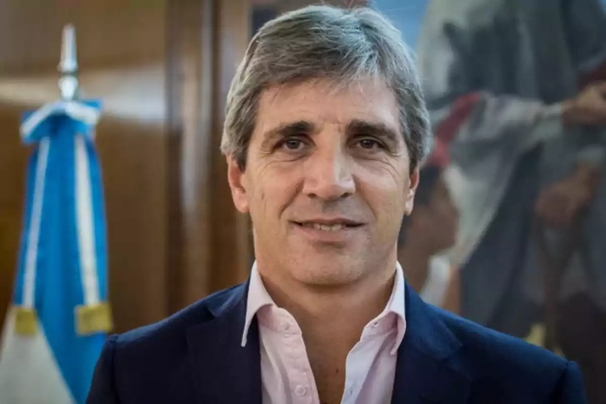 Hombre de cabello canoso con traje oscuro y camisa clara frente a una bandera azul y blanca.