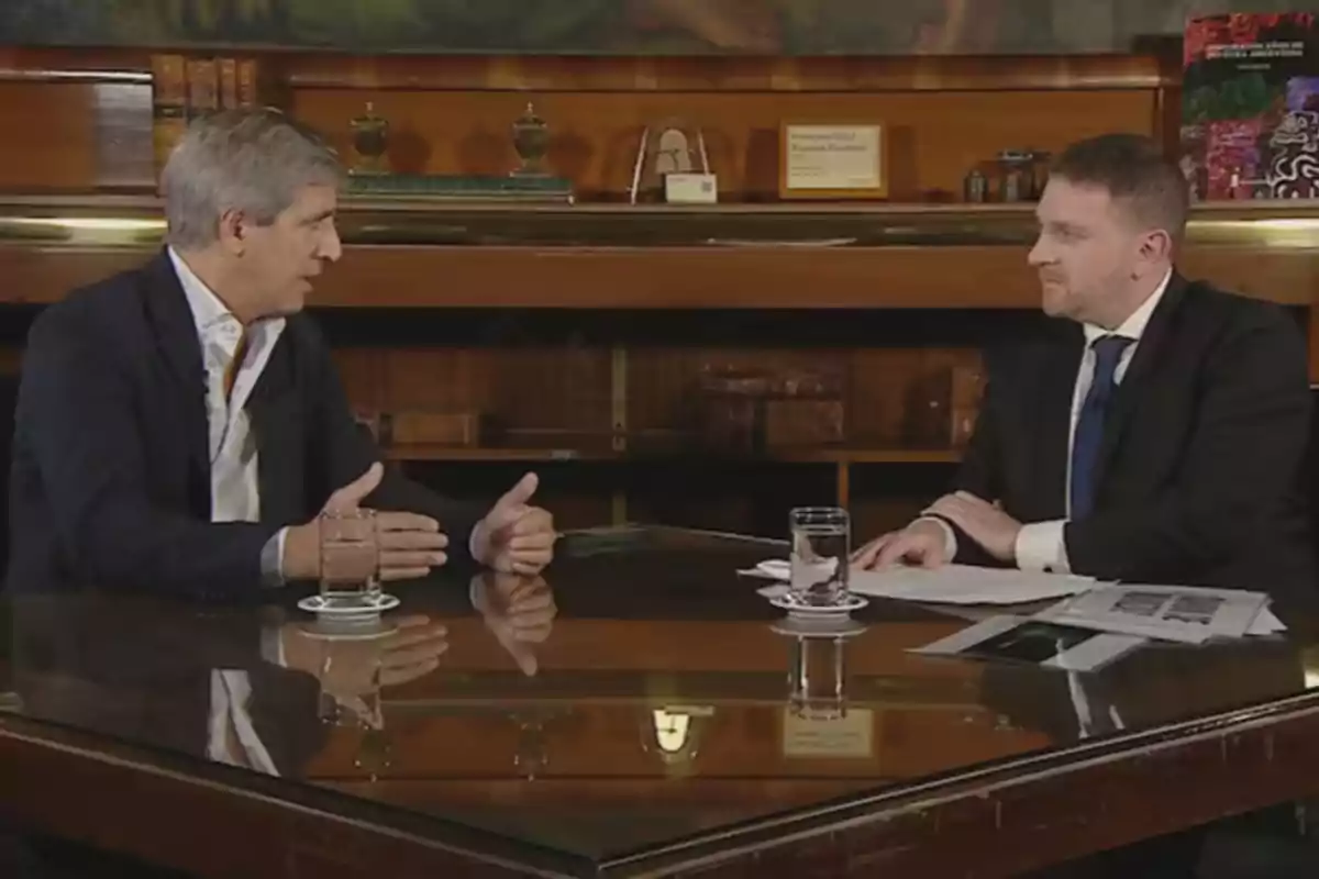 Dos hombres sentados en una mesa de vidrio conversando en un ambiente formal con libros y objetos decorativos en el fondo.