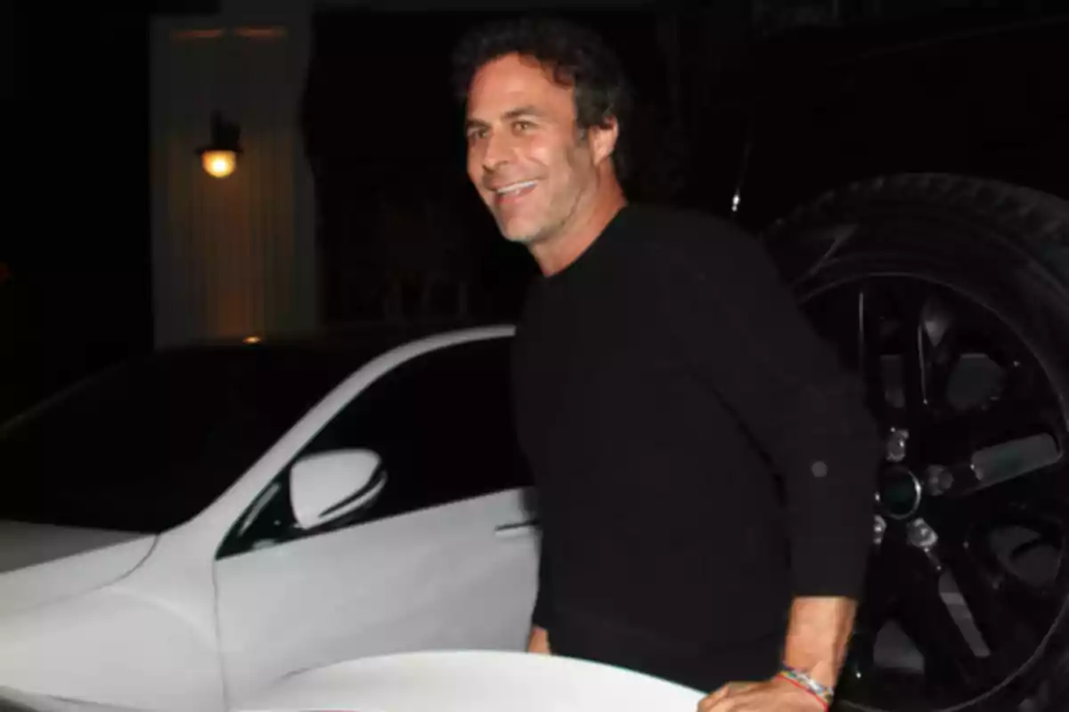 Un hombre sonriente con suéter negro está de pie junto a un automóvil blanco en una escena nocturna. Un hombre sonriente con suéter negro está de pie junto a un automóvil blanco en una escena nocturna.