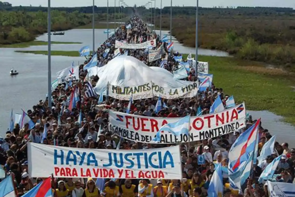 Una multitud de personas marcha sobre un puente sosteniendo pancartas y banderas en un entorno natural con agua y vegetación a los lados.