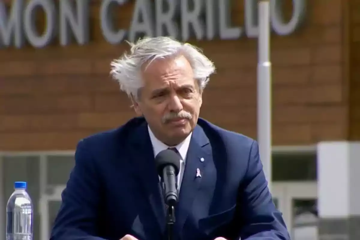 Un hombre de cabello canoso y traje oscuro habla frente a un micrófono con un edificio de fondo.