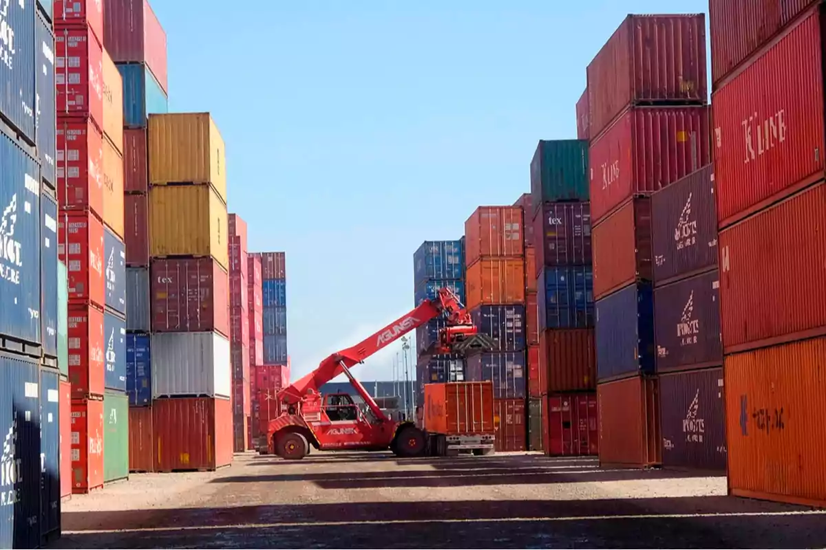 Una grúa roja mueve un contenedor en un puerto lleno de contenedores apilados de varios colores bajo un cielo despejado.