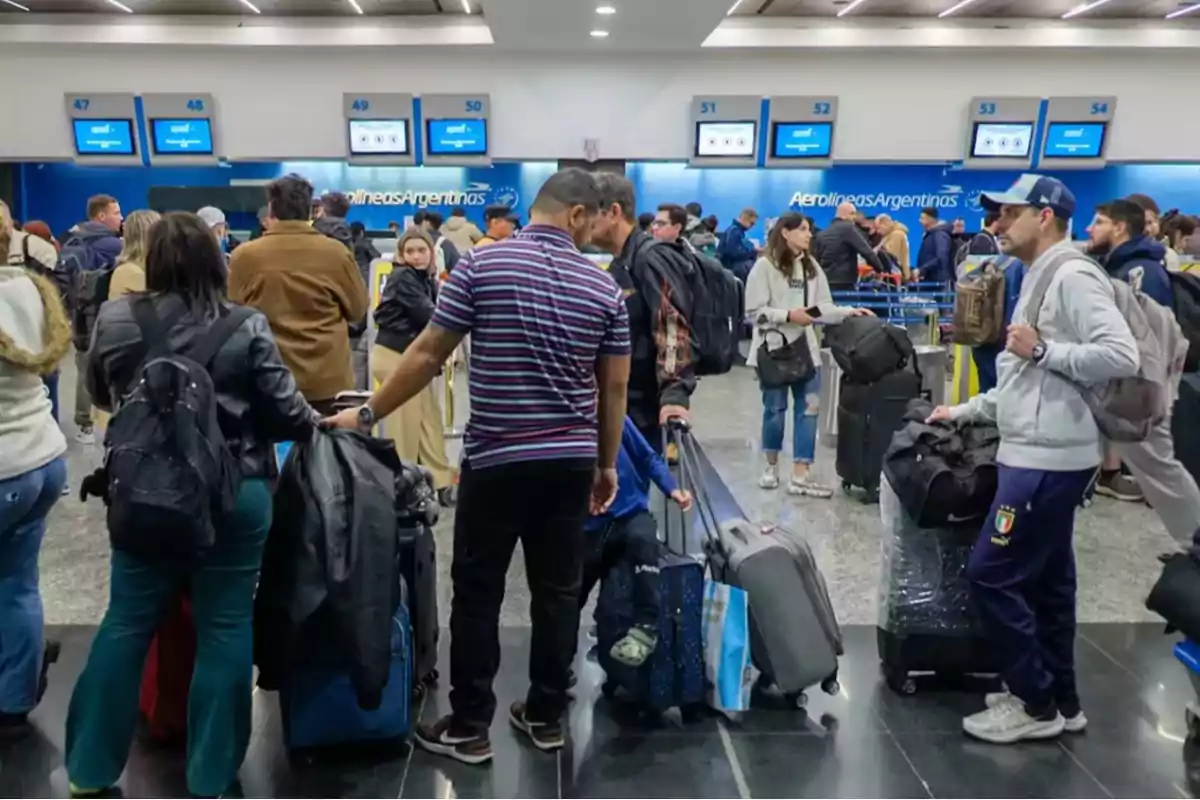 Personas haciendo fila en el área de check-in de un aeropuerto con maletas y pantallas de Aerolíneas Argentinas al fondo.