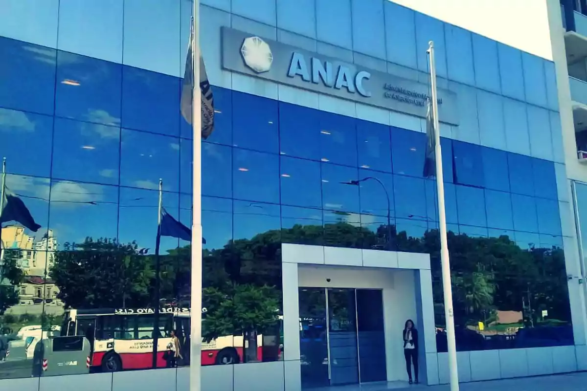 Edificio de la ANAC con fachada de vidrio reflejando el cielo y un autobús en la calle.