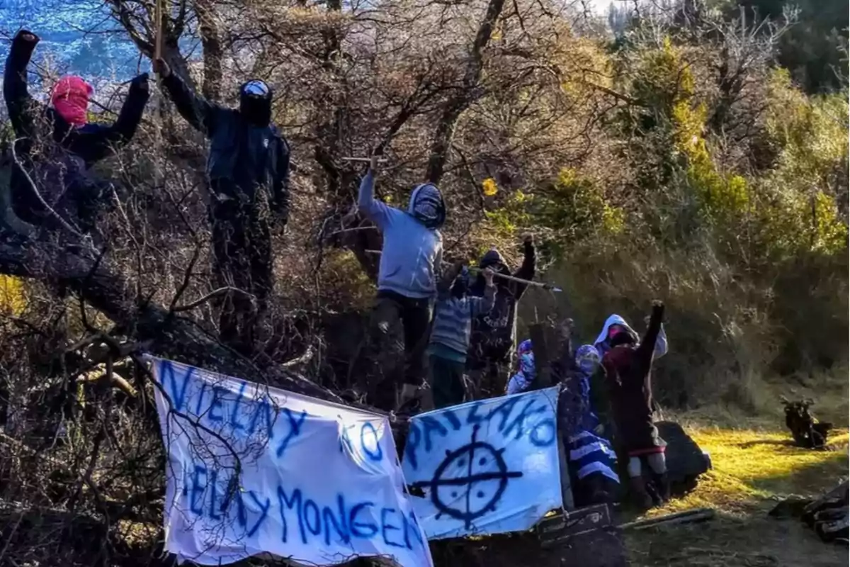 Un grupo de personas con el rostro cubierto levanta los puños y sostiene pancartas en un entorno boscoso.