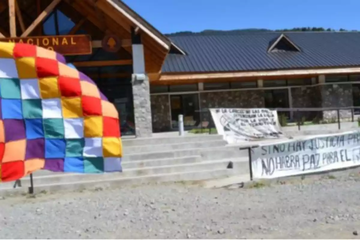 Una bandera multicolor ondea frente a un edificio de madera y piedra con pancartas que contienen mensajes de protesta.