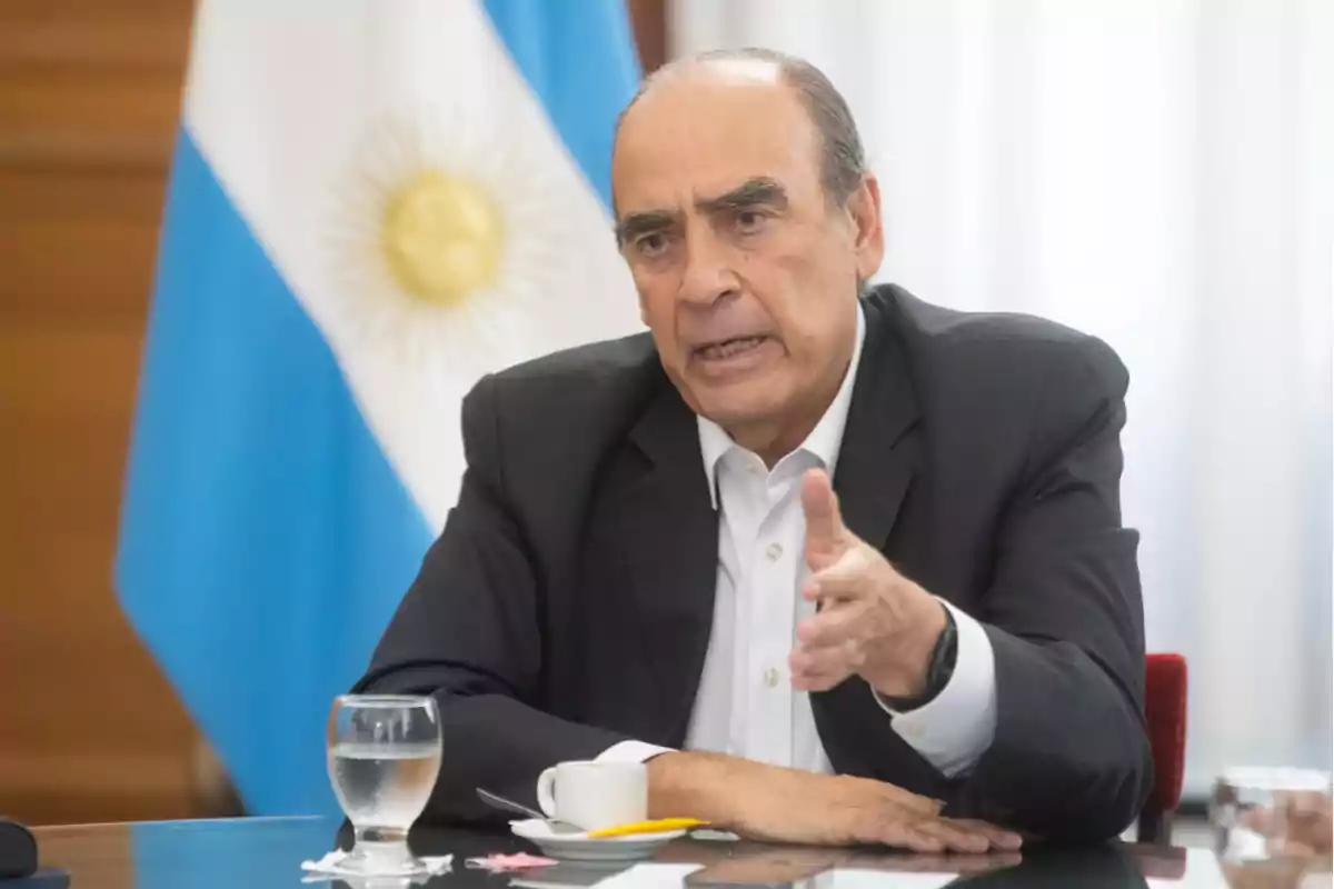 Un hombre de traje oscuro gesticula mientras está sentado en una mesa con una bandera de Argentina de fondo.